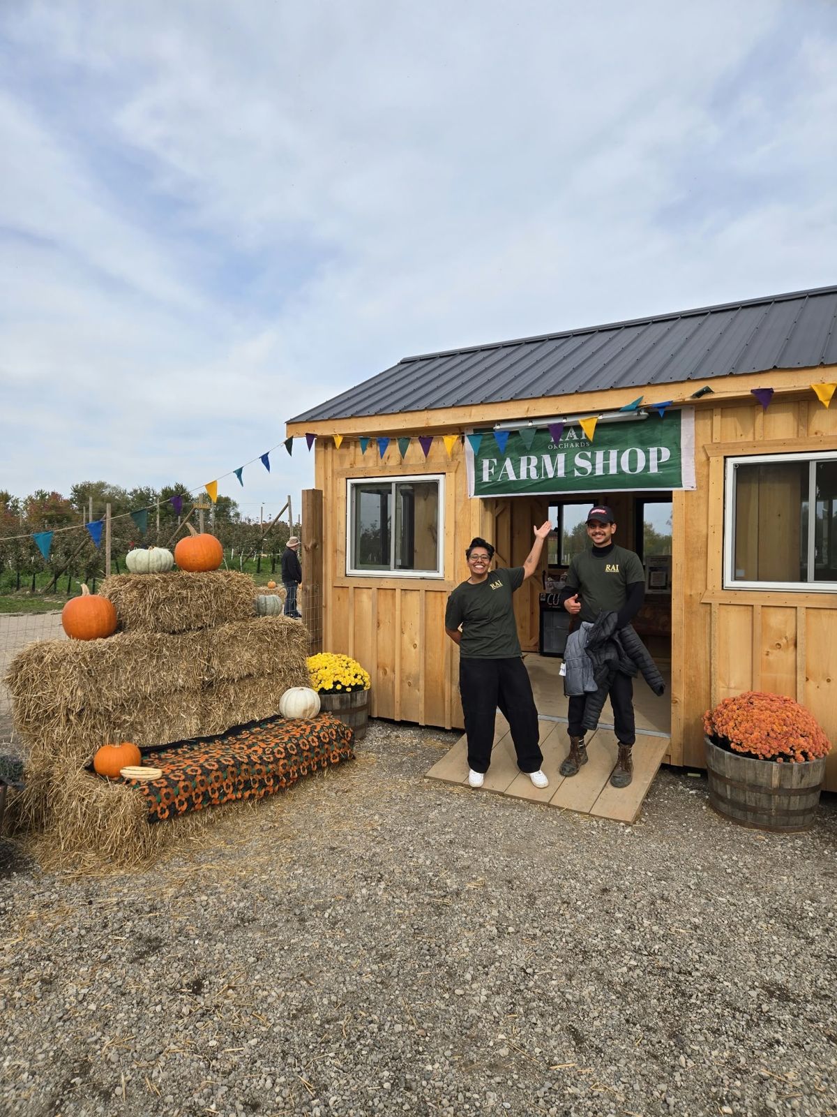 Two members of the Rai Orchards team welcoming visitors outside the farm shop, with pumpkins, hay bales and mums by the door